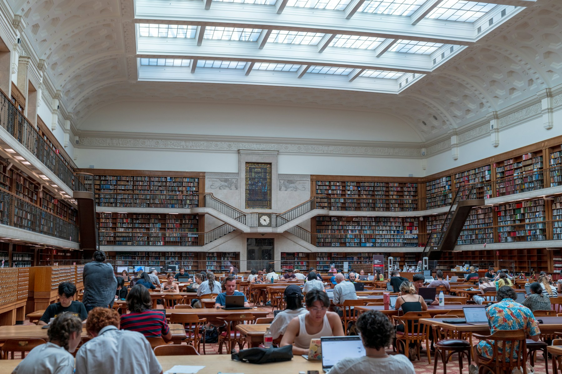 A sunlit library with tall bookshelves and students studying